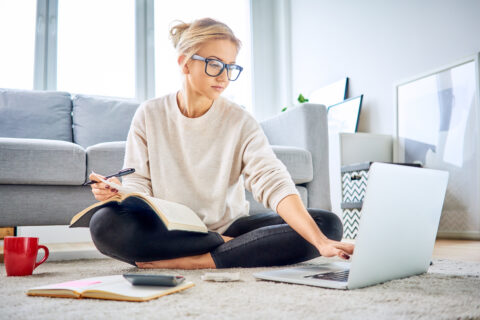 A woman sitting on the floor with a notebook, calculator, and laptop working on a personal budget