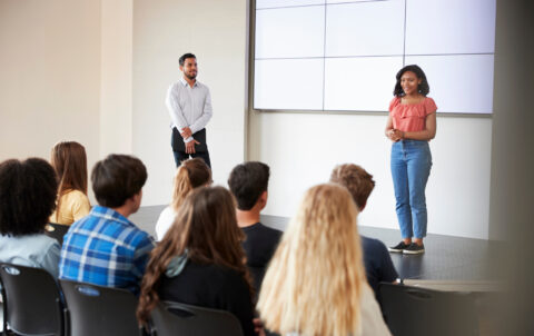 a young woman standing on a platform in front of a screen and people seated while giving a great presentation