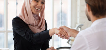A Muslim woman is sitting at a desk shaking a man's hand after a job interview.