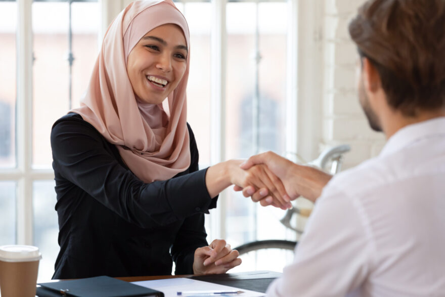 A Muslim woman is sitting at a desk shaking a man's hand after a job interview.
